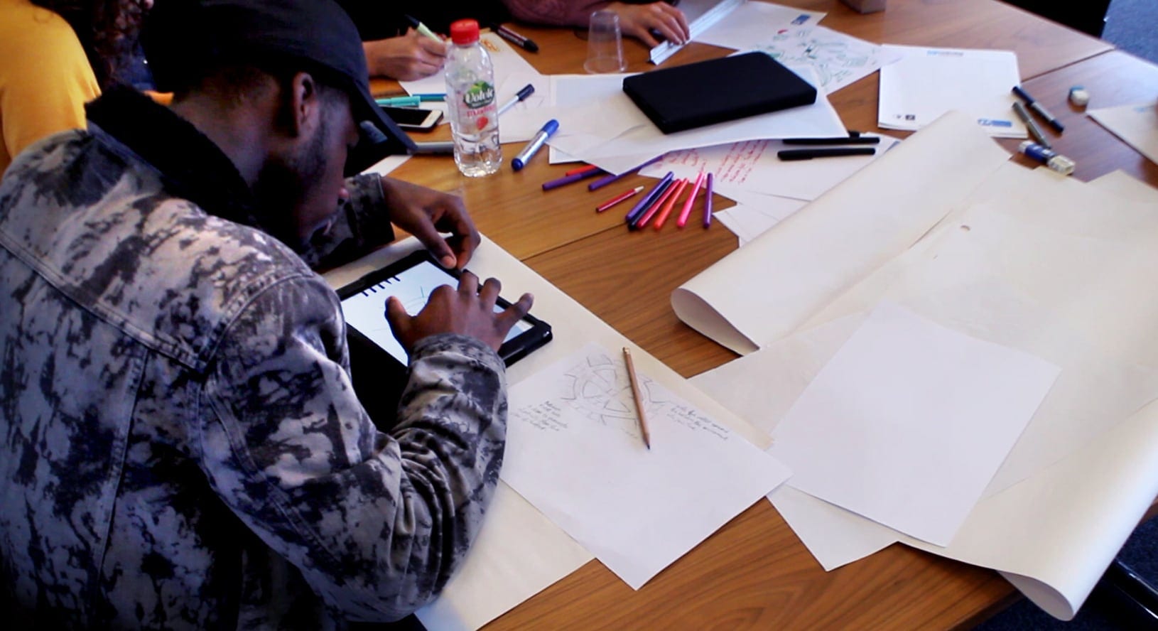 Hands drawing at a table covered in paper and pens