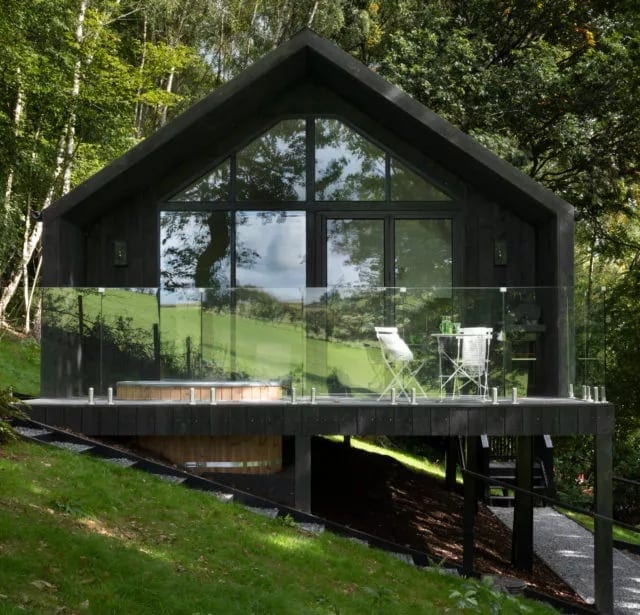 Black timber cabin with a fully glazed gable and glass balcony in woodland